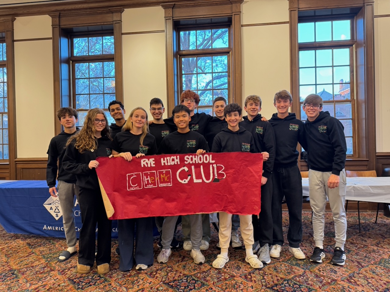 Chemistry Club volunteers holding a Rye High School Chem Club banner at Rye Library.