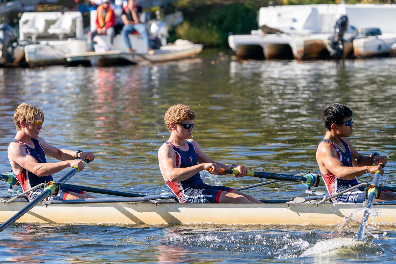 RowAmerica Rye teammates rowing past a dock, with water splashing beside their boat.