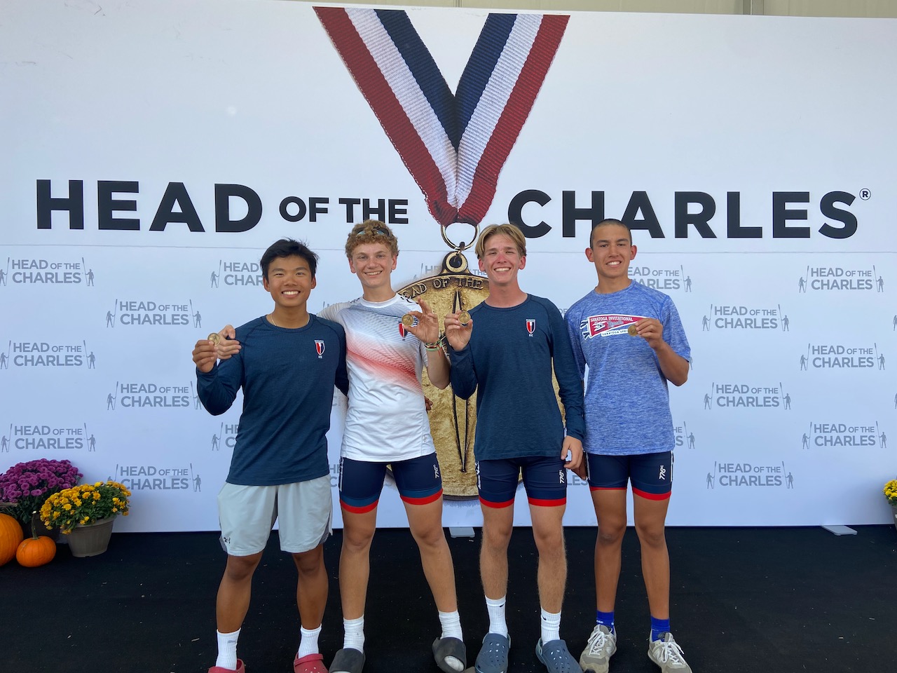 Four RowAmerica Rye teammates holding their medals in front of a Head of the Charles backdrop.